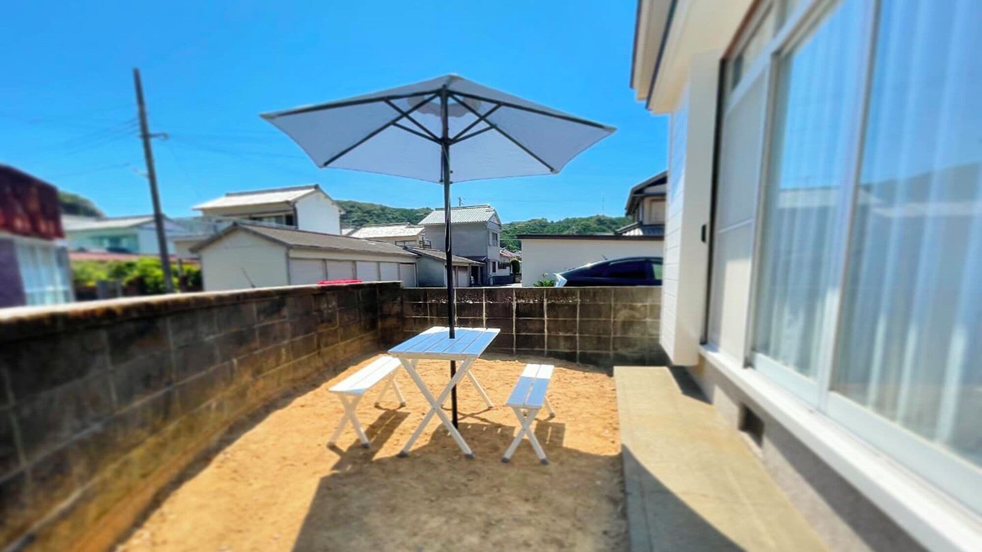A small patio area with a table and two chairs, a white umbrella, and a view of houses in the background.