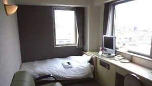 Habitación de hotel pequeña con una cama individual, un escritorio con computadora y teléfono, una silla y un ventanal con cortinas.