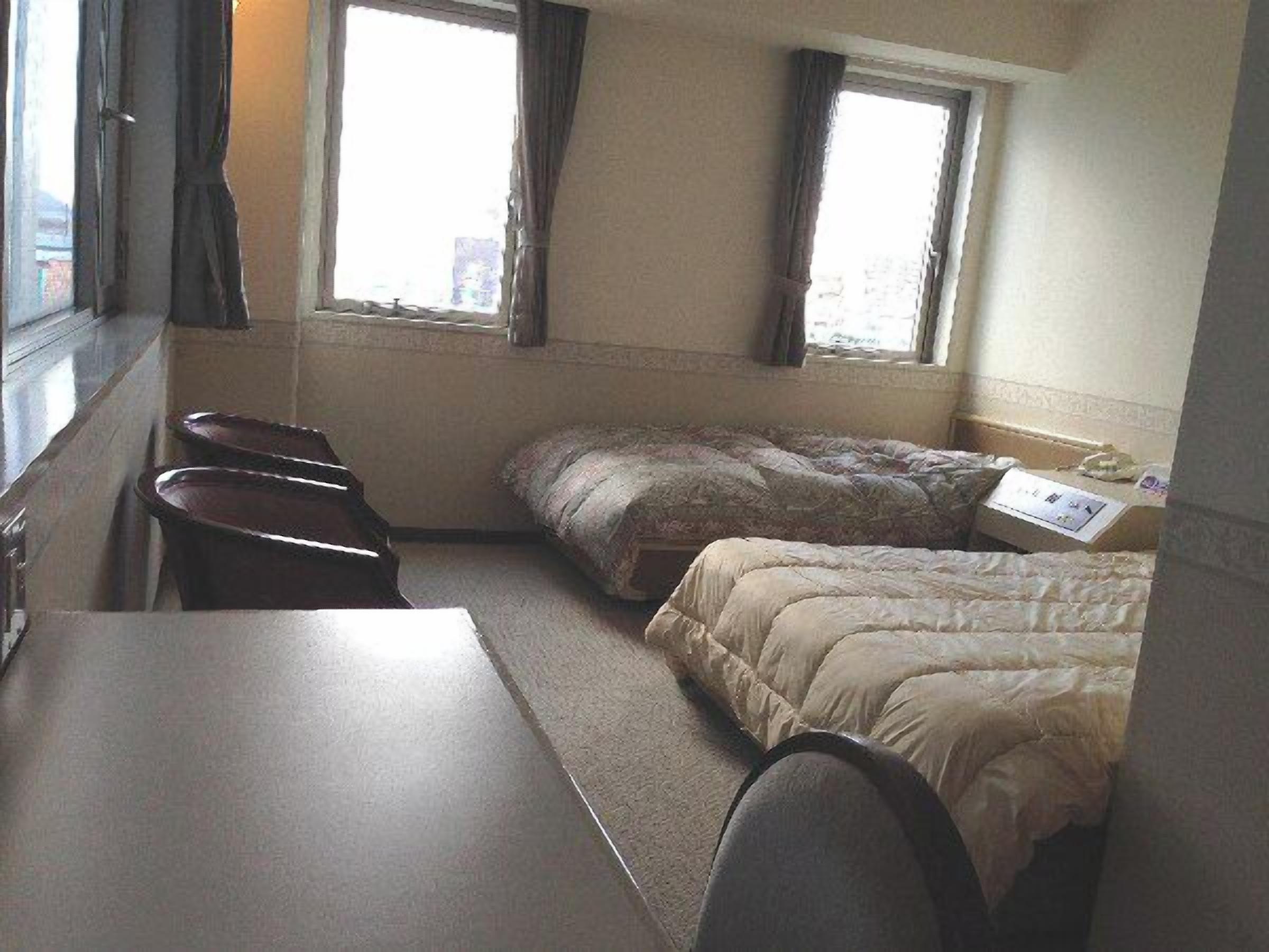 Ein kleines, kompaktes Zimmer mit einem Einzelbett, einem Schreibtisch und einem Stuhl. Zwei Fenster mit Vorhängen und ein kleiner Tisch mit Papier darauf.