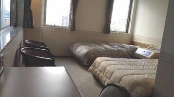 A small, compact room with a single bed, a desk, and a chair. There are two windows with curtains, and a small table with a paper on it.