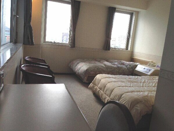 A small, compact room with a single bed, a desk, and a chair. There are two windows with curtains, and a small table with a paper on it.