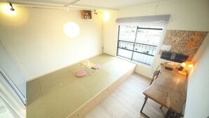Una habitación con zona de estar con estera de tatami, un escritorio de madera, una ventana con vistas y una estantería montada en la pared.