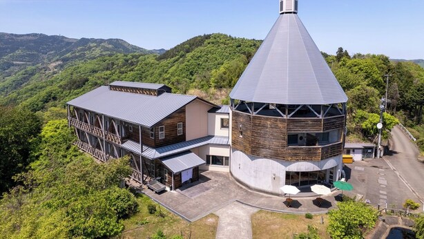 Ein Gebäude mit einem Kegelturm, eine Holzkonstruktion mit Metallbedachung und eine gepflasterte Fläche mit Außenplätzen.