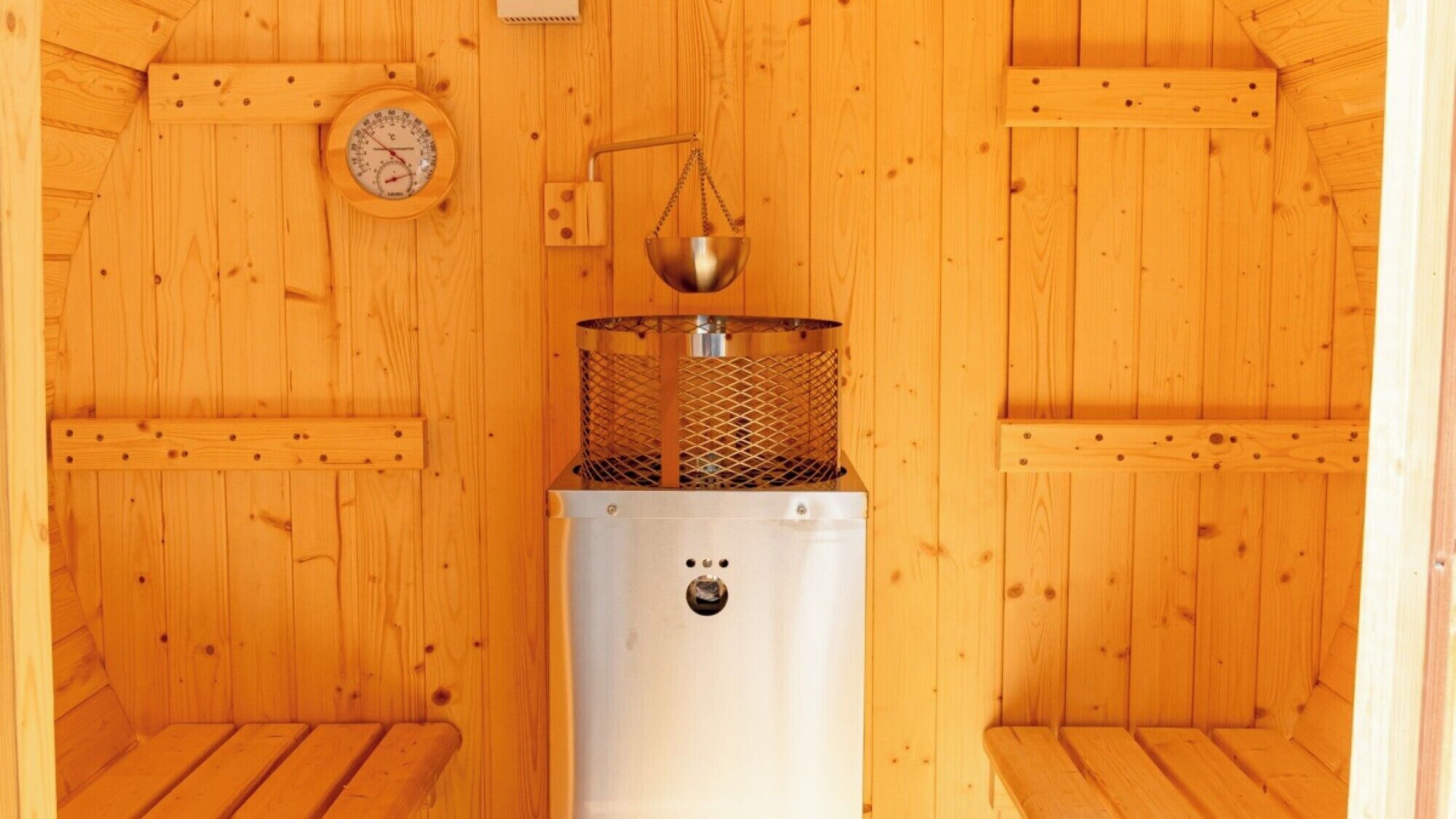 Una sala de sauna con paredes de madera, un calentador de sauna, un termómetro y un recipiente metálico.