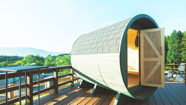 A wooden barrel-shaped structure with an open door, a wooden deck, and a view of a lake and mountains.