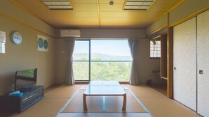 A room with tatami flooring, a low table, a TV on a cabinet, a large window with curtains, and a sliding door.