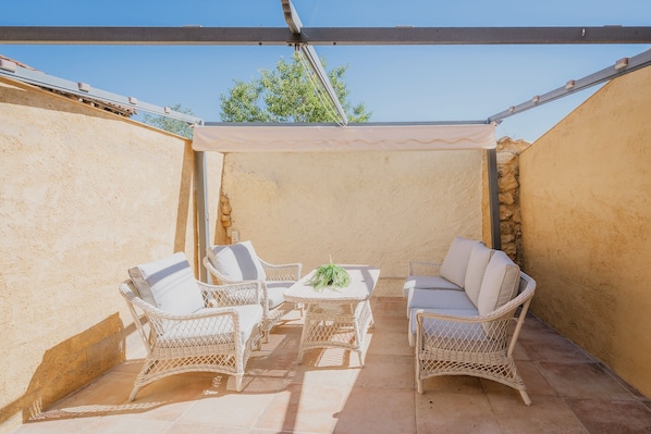 A covered patio area with wicker furniture and a table, featuring a stone wall and a clear blue sky.