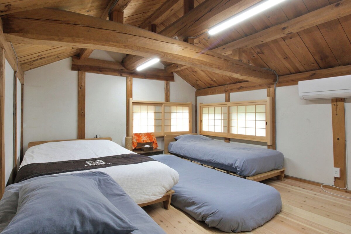 A room with a wooden ceiling, a bed with a white mattress, two single beds with grey bedding, a small table with a lamp, and a window with wooden frames.