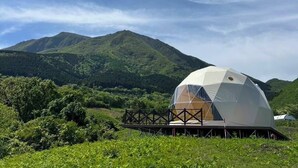 A geodesic dome structure with a wooden deck in a grassy field, surrounded by hills and a clear sky. - Resonate Glamping Hoshi no Oka (Taketa)