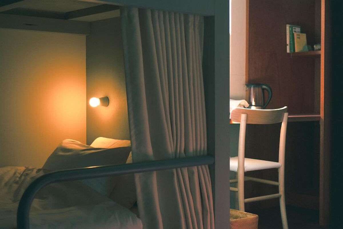 Una cama individual con mesita de noche y silla, una lámpara de pared, y una estantería con libros y una tetera.