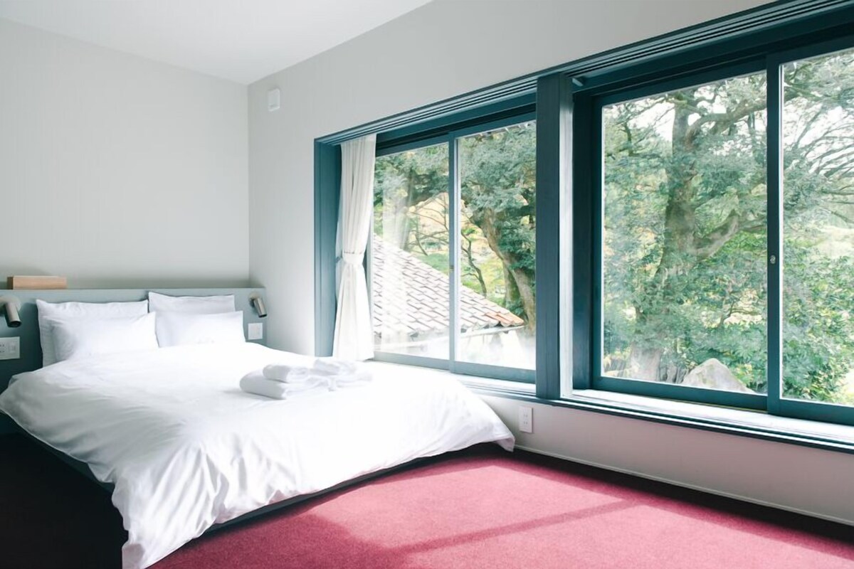 Habitación de hotel con una cama grande, una mesita de noche, una ventana con cortinas y vistas a árboles.