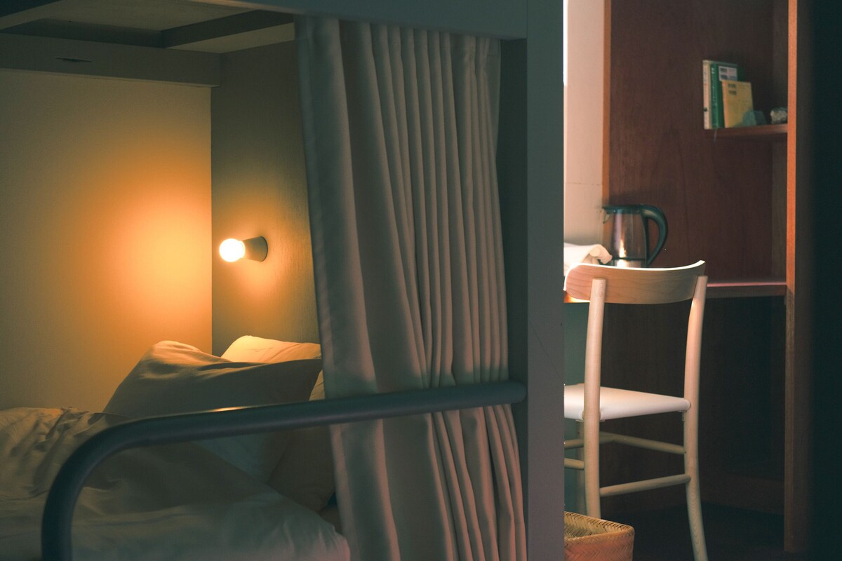 Una habitación con cama, una silla, una mesa con tetera y una estantería.