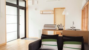 Een moderne woonkamer met een houten tafel, een zwarte bank met gestreepte kussens, een keuken met een spoelbak en fornuis, en een schuifdeur van glas die uitkomt op een balkon.