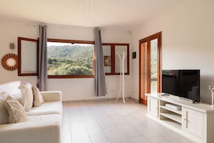 Un salón con un sofá, un televisor, un mueble bajo y vistas a un paisaje verde a través de una ventana.