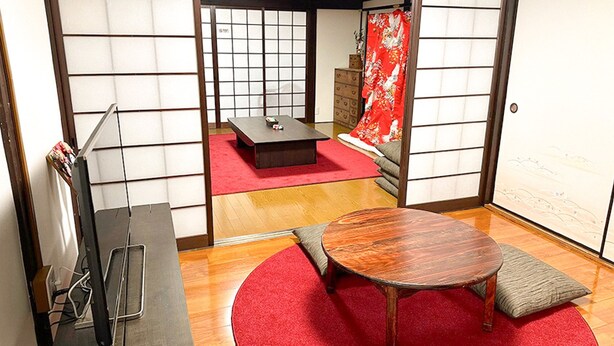 A traditional Japanese room with tatami flooring, a low table with cushions, a television, and a sliding door with a red curtain.