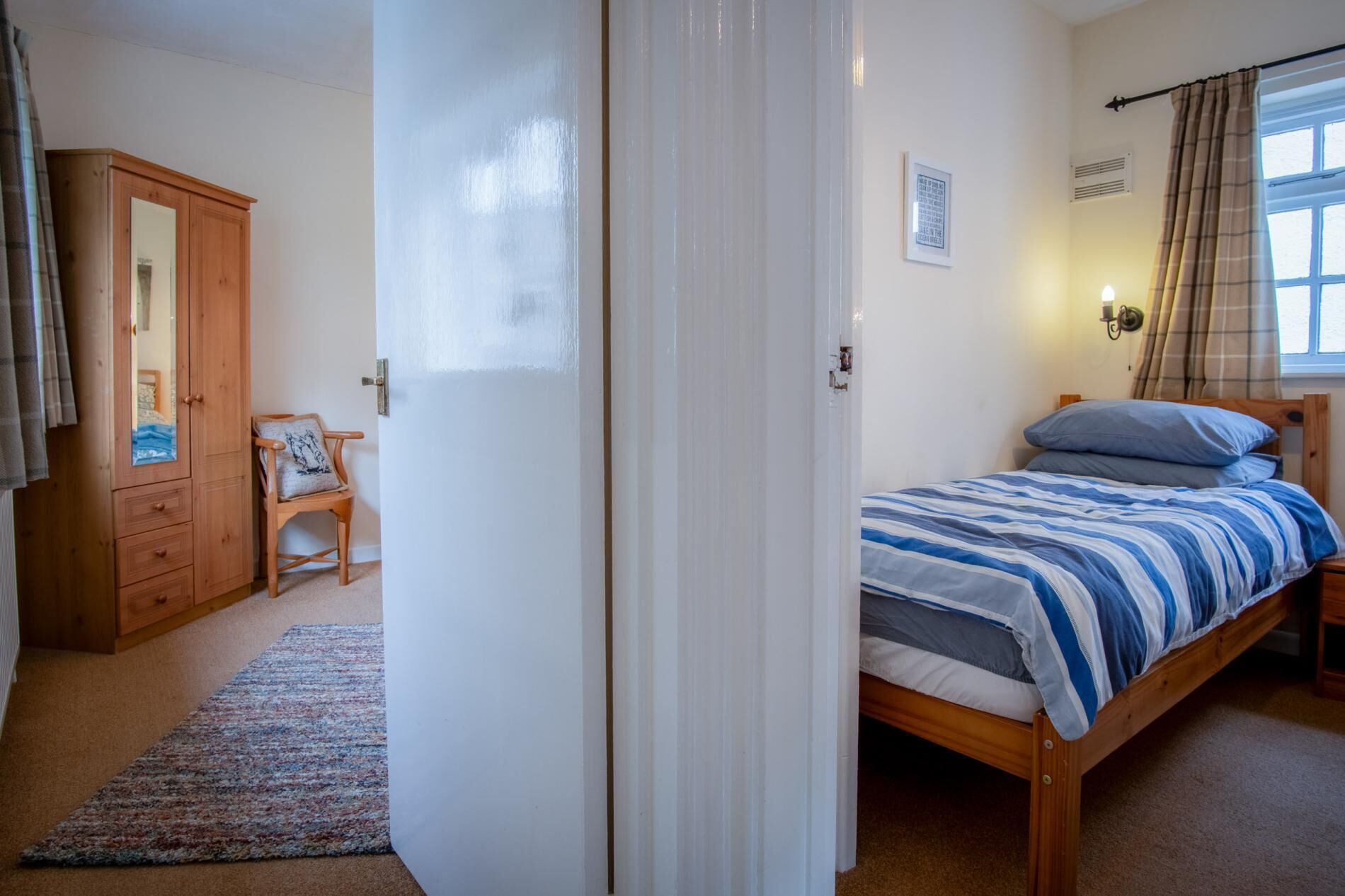 A bedroom with a bed, a wooden wardrobe, a chair, a rug, and a window with curtains.