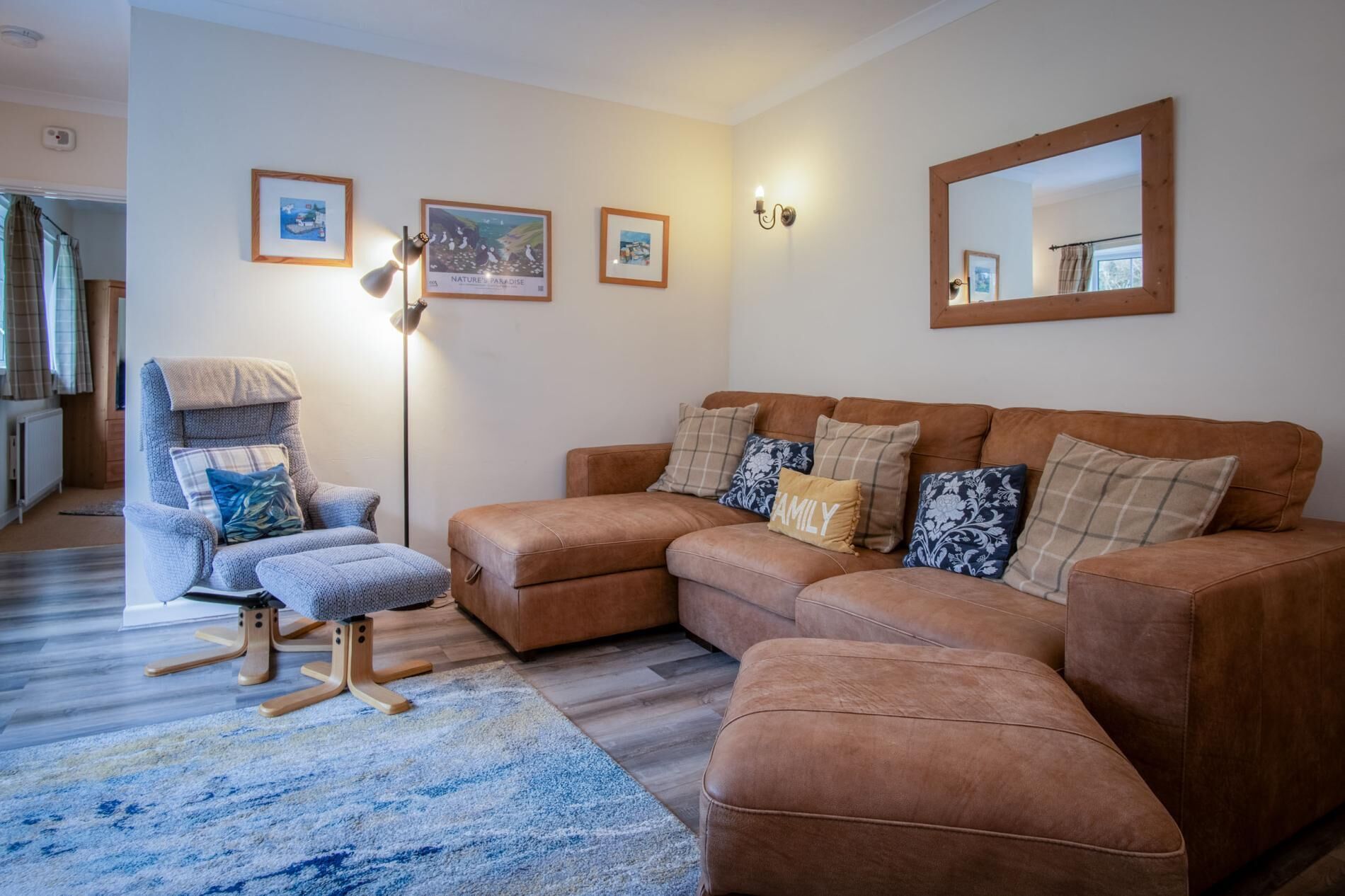 Una sala de estar con un sofá seccional marrón, un sillón gris y un tapete azul estampado. Hay tres cuadros en la pared y una lámpara de pie.