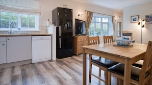 A modern kitchen with a dishwasher, refrigerator, and a dining area with a table and chairs. - Garden Cottage - 2 Bedroom Cottage - Penally (Tenby)