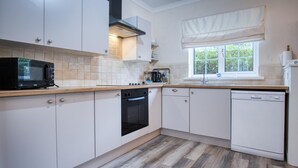 A modern kitchen with white cabinets, a black microwave, a dishwasher, and a window with a white valance. - Garden Cottage - 2 Bedroom Cottage - Penally (Tenby)