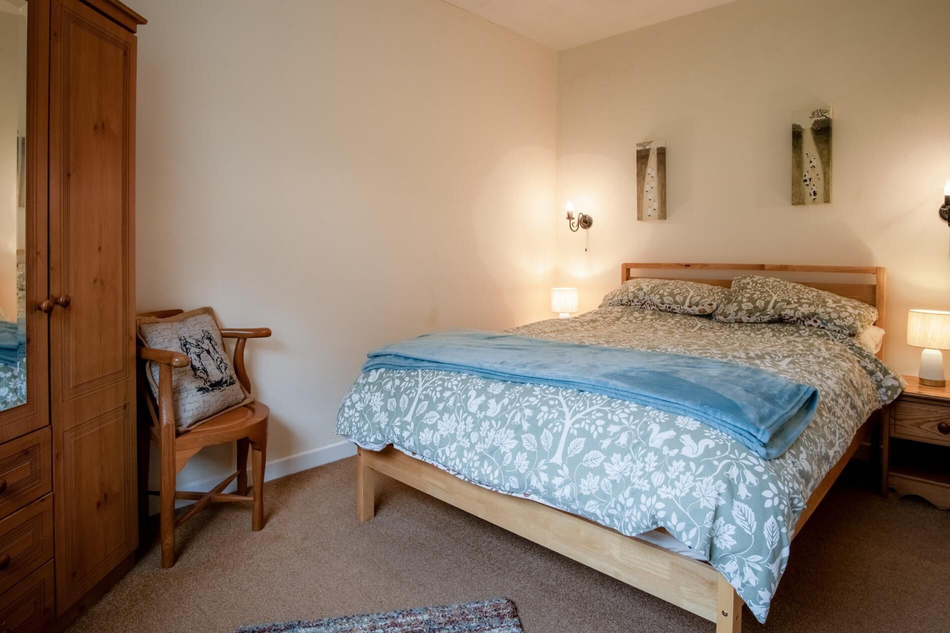 Een slaapkamer met een houten bed, een nachtkastje met een lamp, een houten garderobe en een stoel met een kussen.