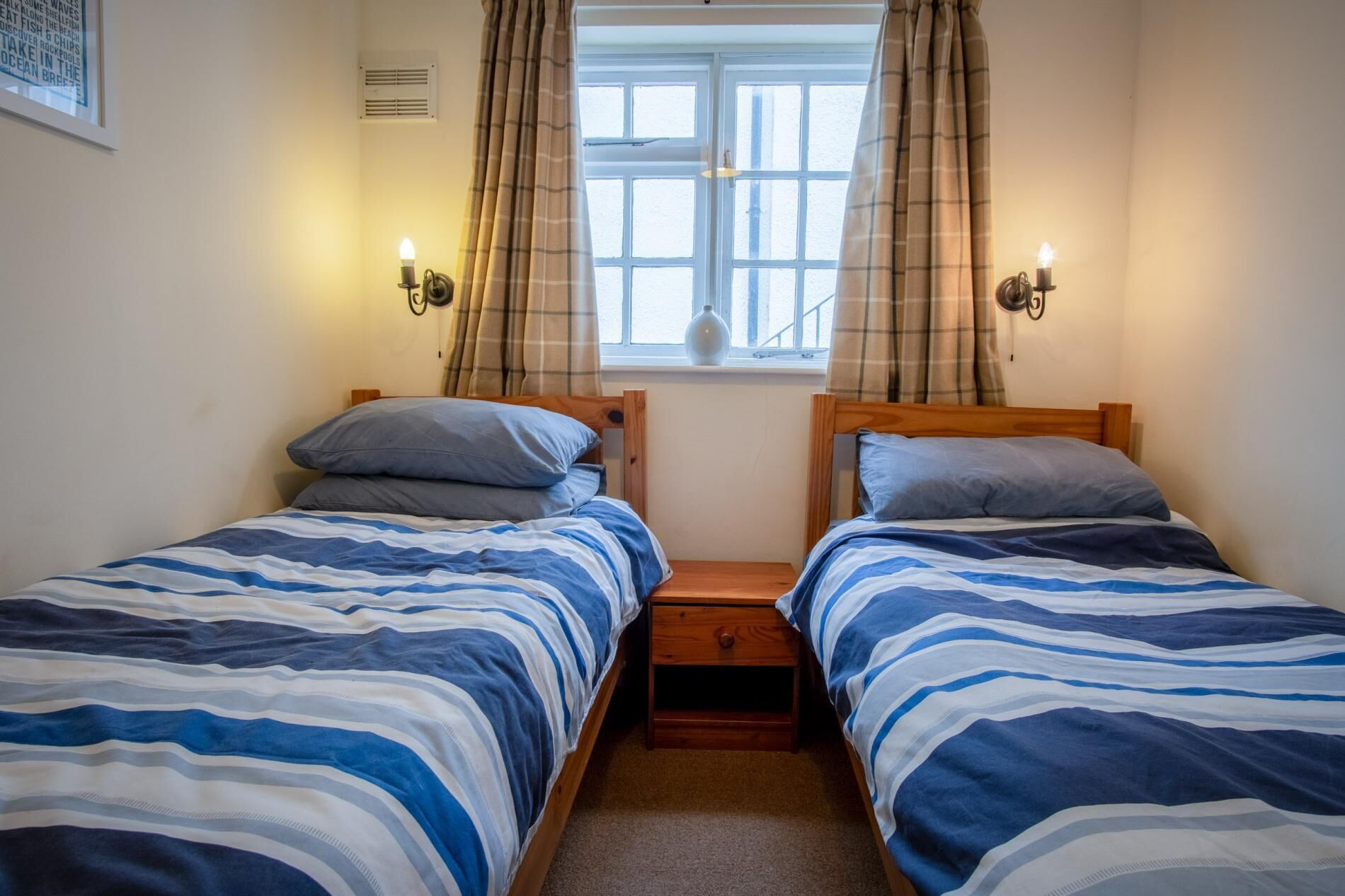 A bedroom with two single beds, each with a wooden headboard, a small bedside table, and a lamp. There is a window with curtains and a view of the outside.