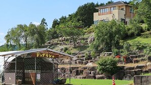 A rustic outdoor seating area with a covered structure, large rocks, and a distant building on a hillside. - Kappo Ryokan Magohachi (Nakatsugawa)