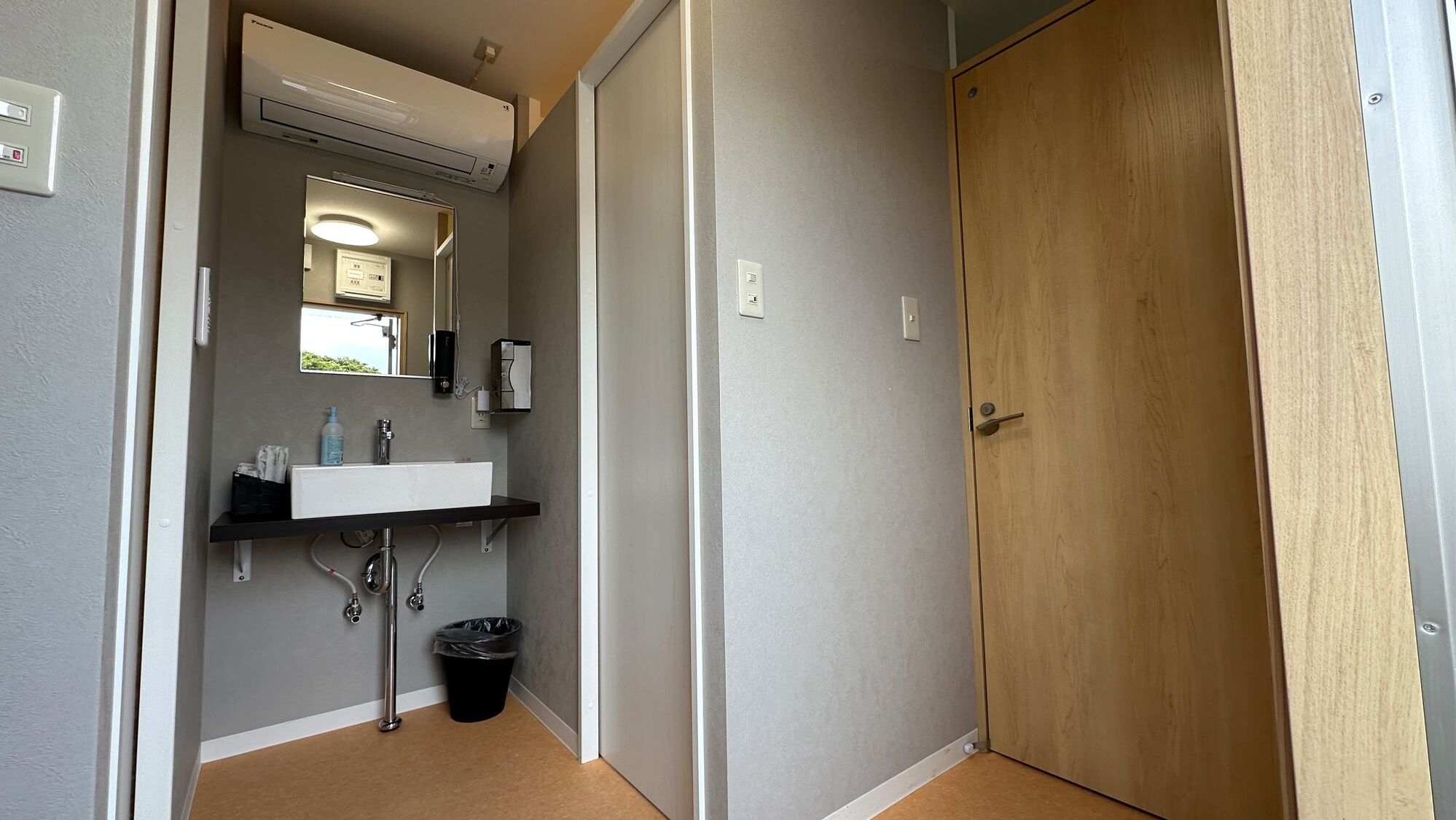 A narrow hallway with a wooden door, a white sink with a mirror, and a trash bin.