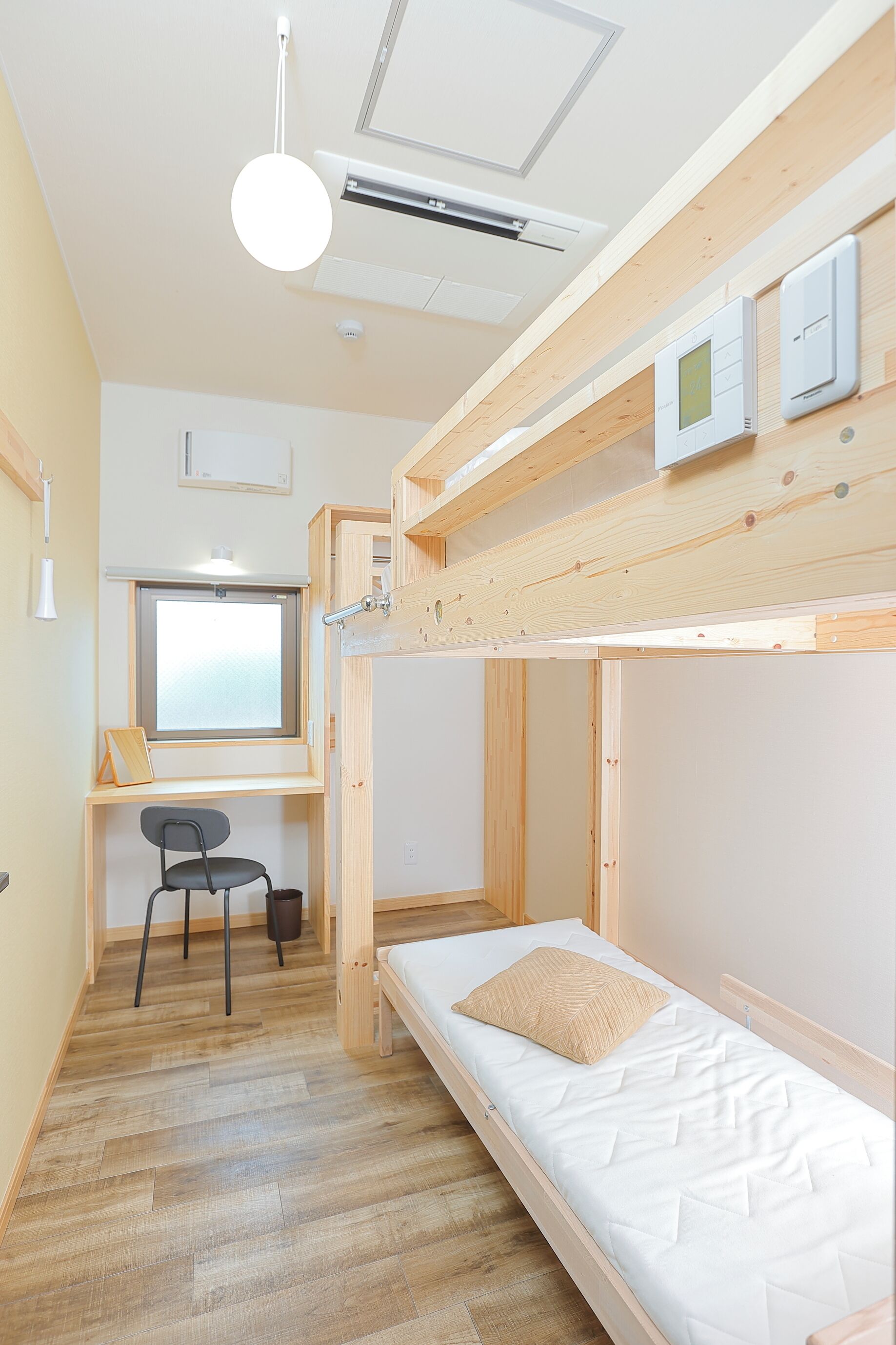 A compact room with a bunk bed, a desk with a chair, a window, and a ceiling light fixture.