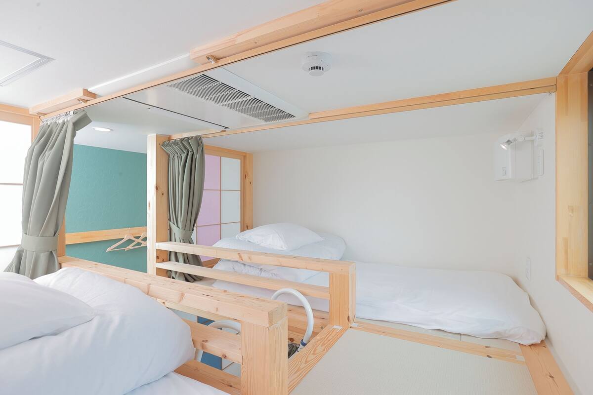 Une chambre avec un lit superposé en bois, une fenêtre avec des rideaux, un climatiseur fixé au mur et un petit miroir.