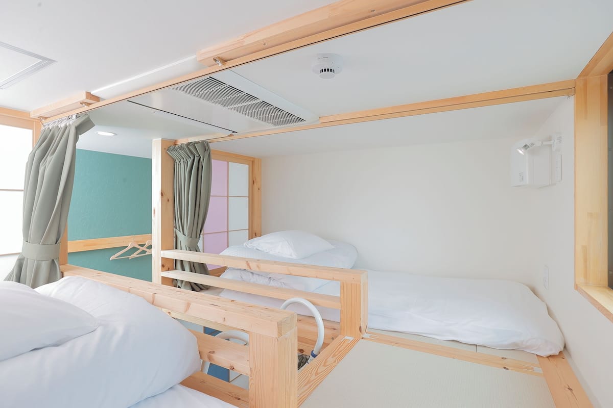 Une chambre avec un lit superposé en bois, une fenêtre avec des rideaux, un climatiseur fixé au mur et un petit miroir.