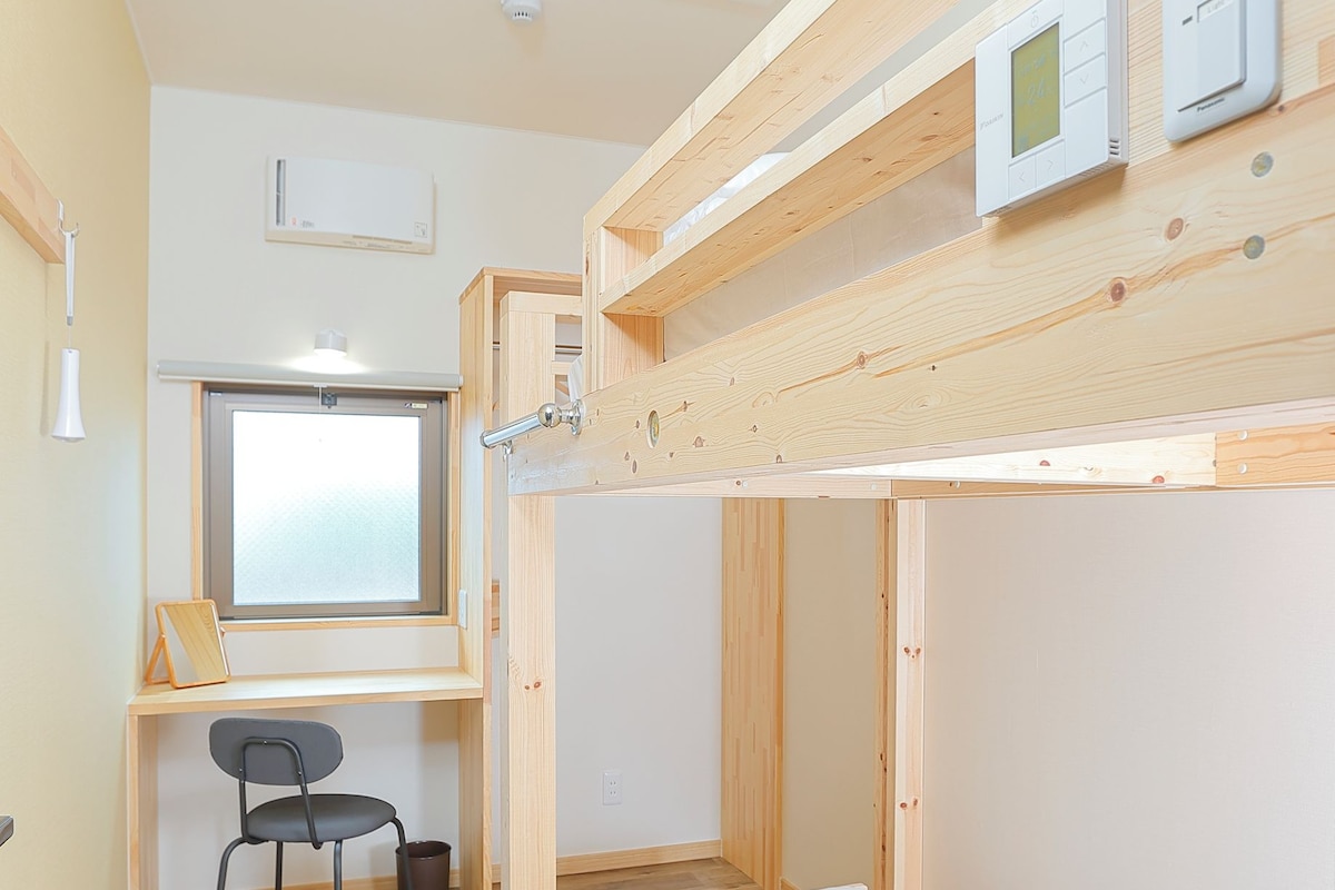 A compact room with a bunk bed, a desk with a chair, a window, and a ceiling light fixture.