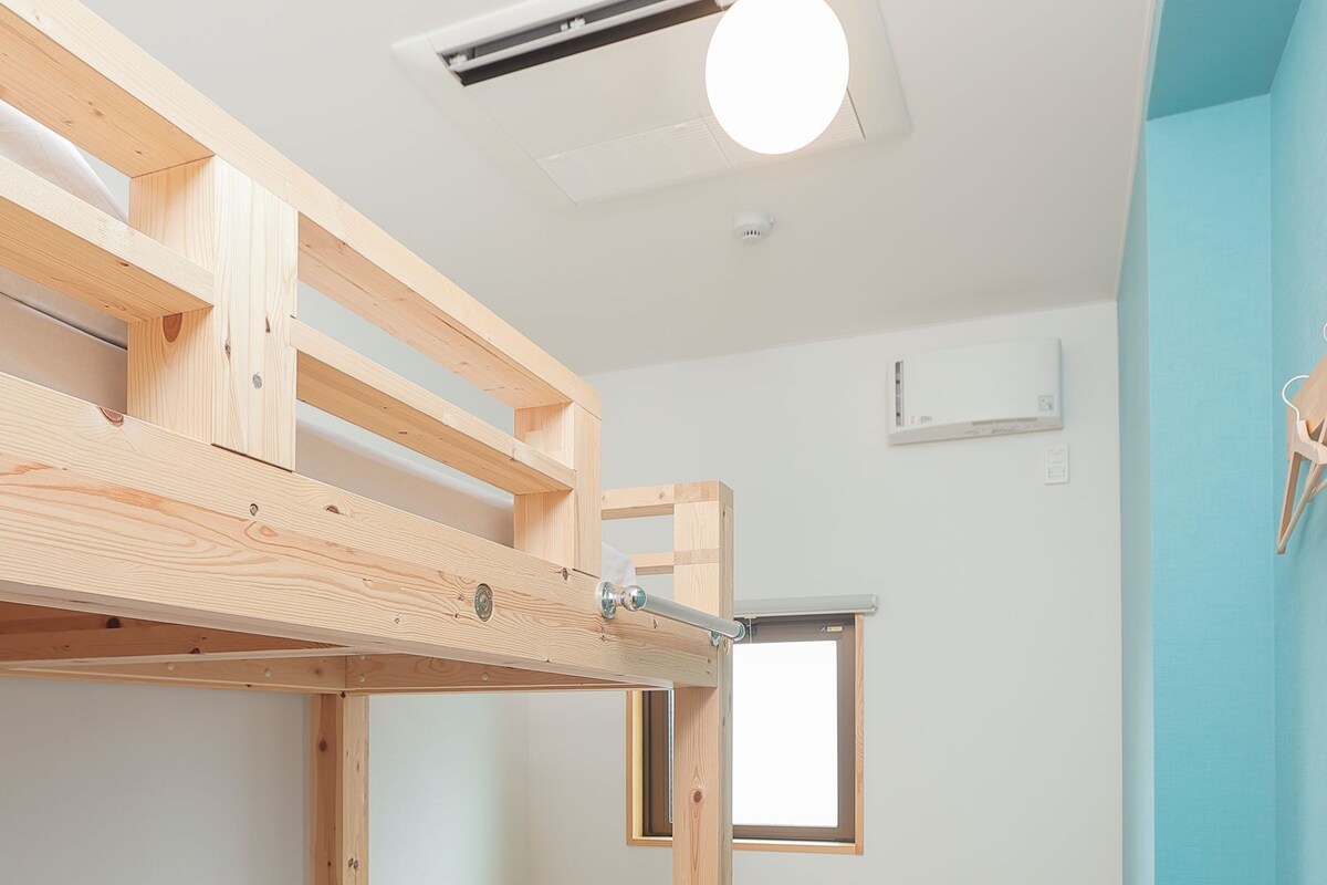 Un lit superposé avec une structure en bois, une petite table, une fenêtre et un luminaire fixé au plafond.