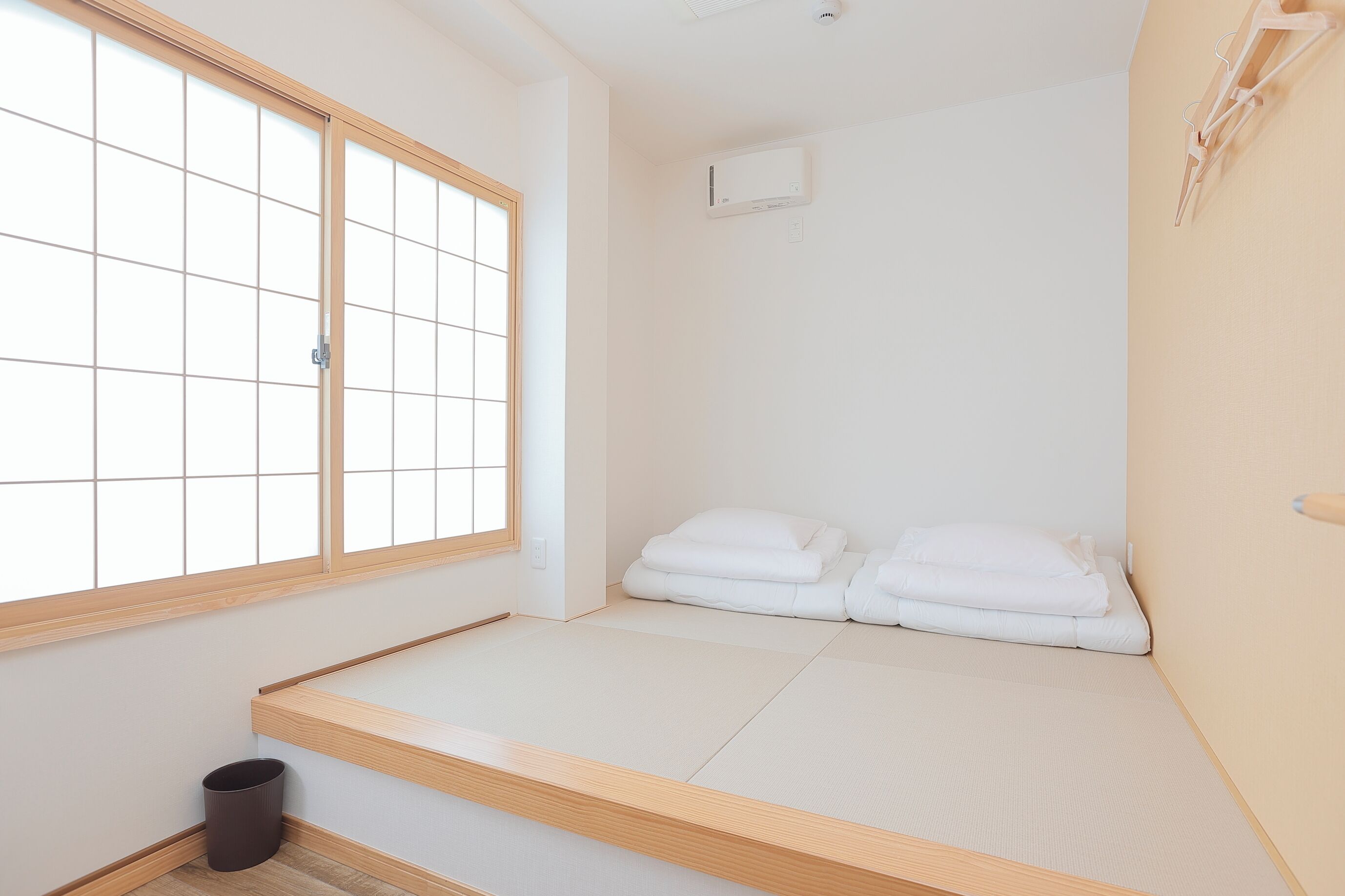 A minimalist room with tatami flooring, a futon bed, a window with a grid pattern, and a wall-mounted air conditioner.