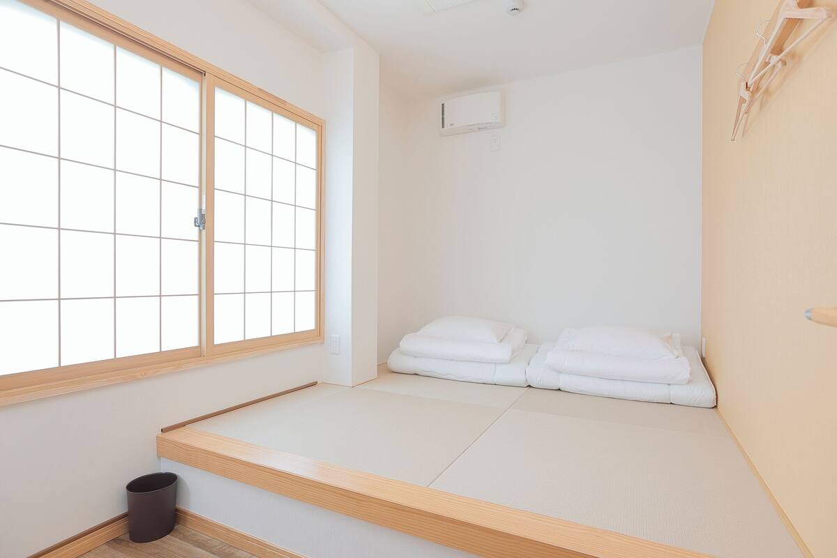 Une pièce minimaliste avec un sol recouvert de tatamis, un futon servant de lit, une fenêtre à carreaux et un climatiseur fixé au mur.
