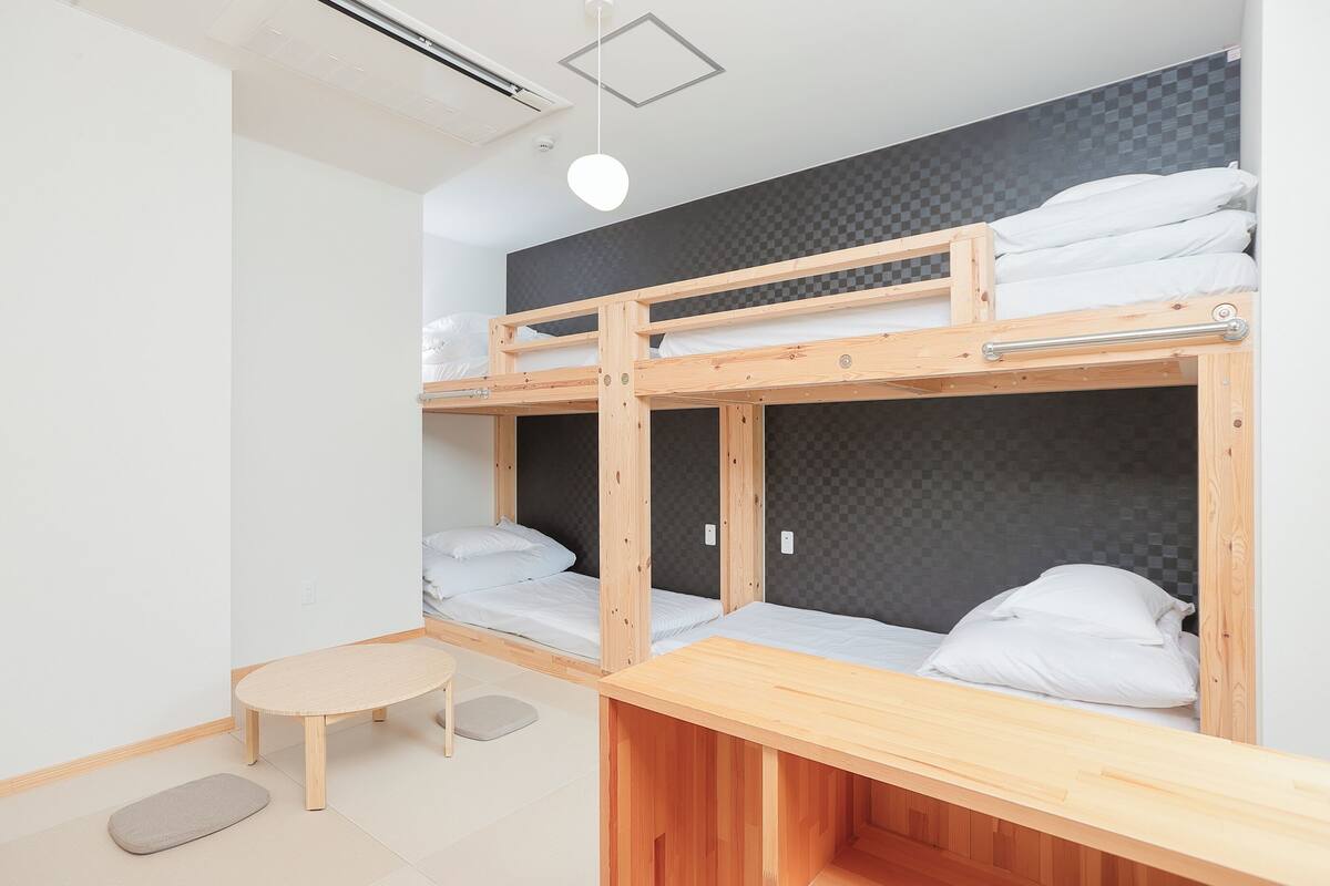 Une chambre avec un lit superposé, une petite table et un banc, un luminaire au plafond et un mur à carreaux.