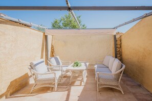 A covered patio area with wicker furniture and a table, featuring a stone wall and a clear blue sky.