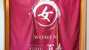 Entrada de un hotel con un cartel rojo que muestra el logotipo y el nombre 'WOMEN YUJU'.