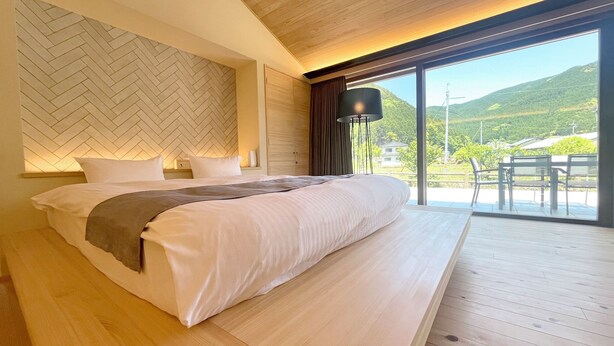A bedroom with a bed, a nightstand, a lamp, and a view of a balcony with chairs. - Centennial Forest Hotel Shiori (Nishiawakura)