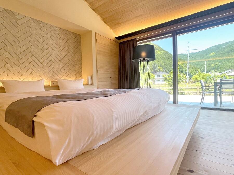 Un dormitorio con cama, mesita de noche, lámpara y vistas a un balcón con sillas.