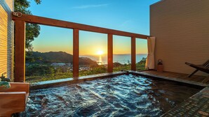 Una vasca idromassaggio con un'area prendisole in legno, affacciata su un panorama mozzafiato dell'oceano e delle colline al tramonto.