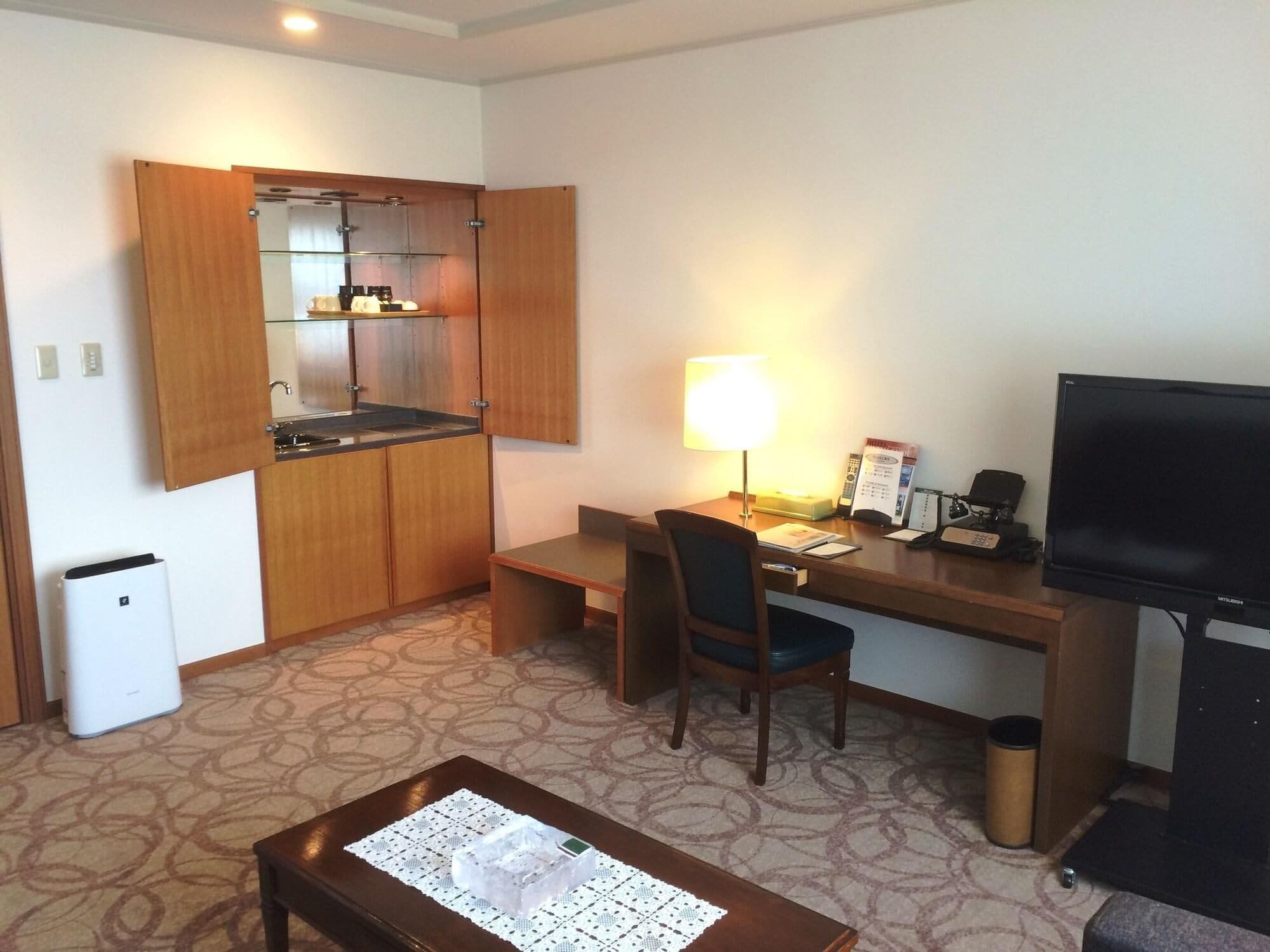 A compact hotel room with a kitchenette, a desk with a chair, a television, and a trash bin.