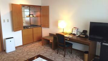 A compact hotel room with a kitchenette, a desk with a chair, a television, and a trash bin.