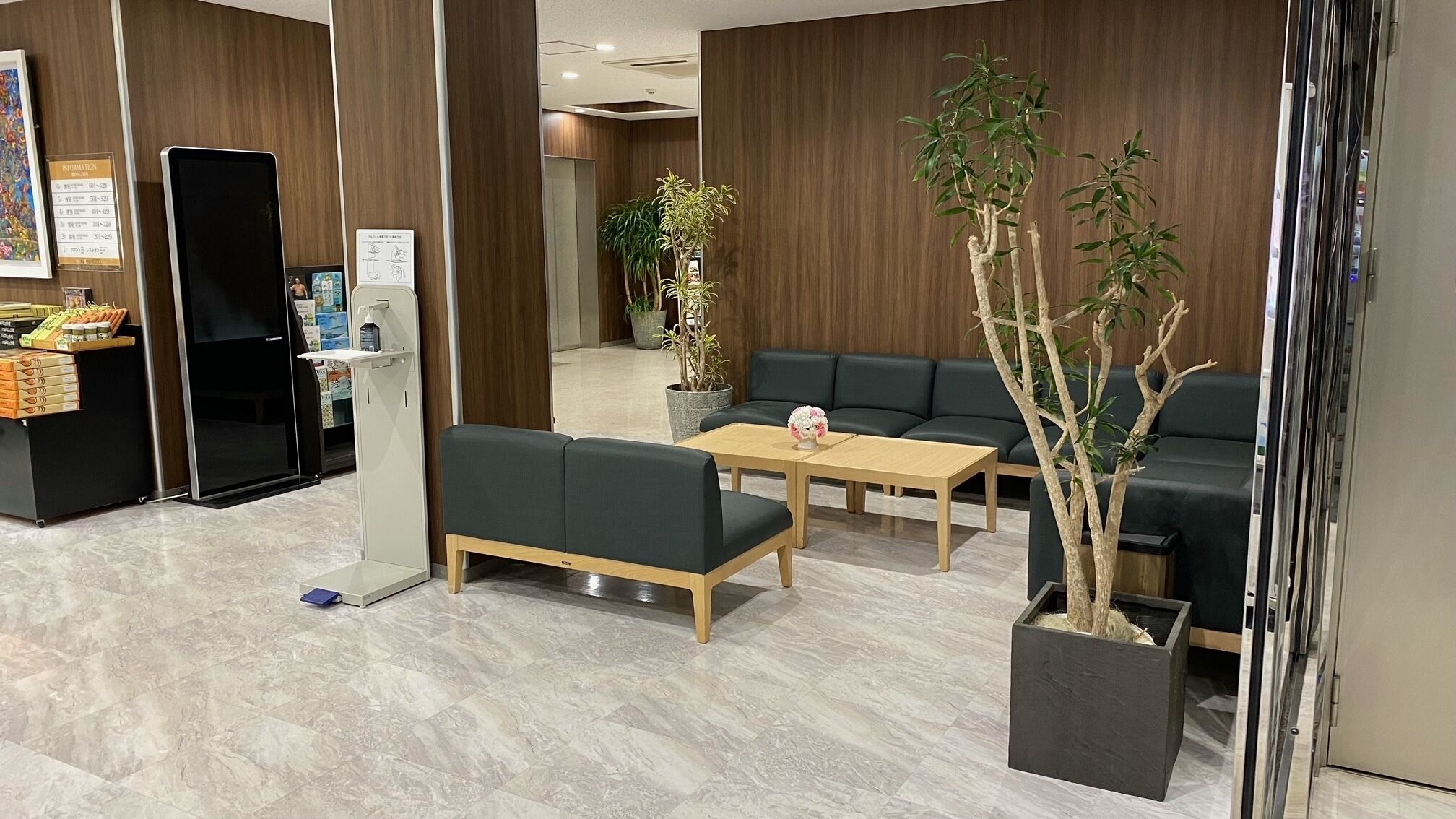 A modern reception area with a sofa, a coffee table, and a reception desk. There are potted plants and a wall-mounted information board.