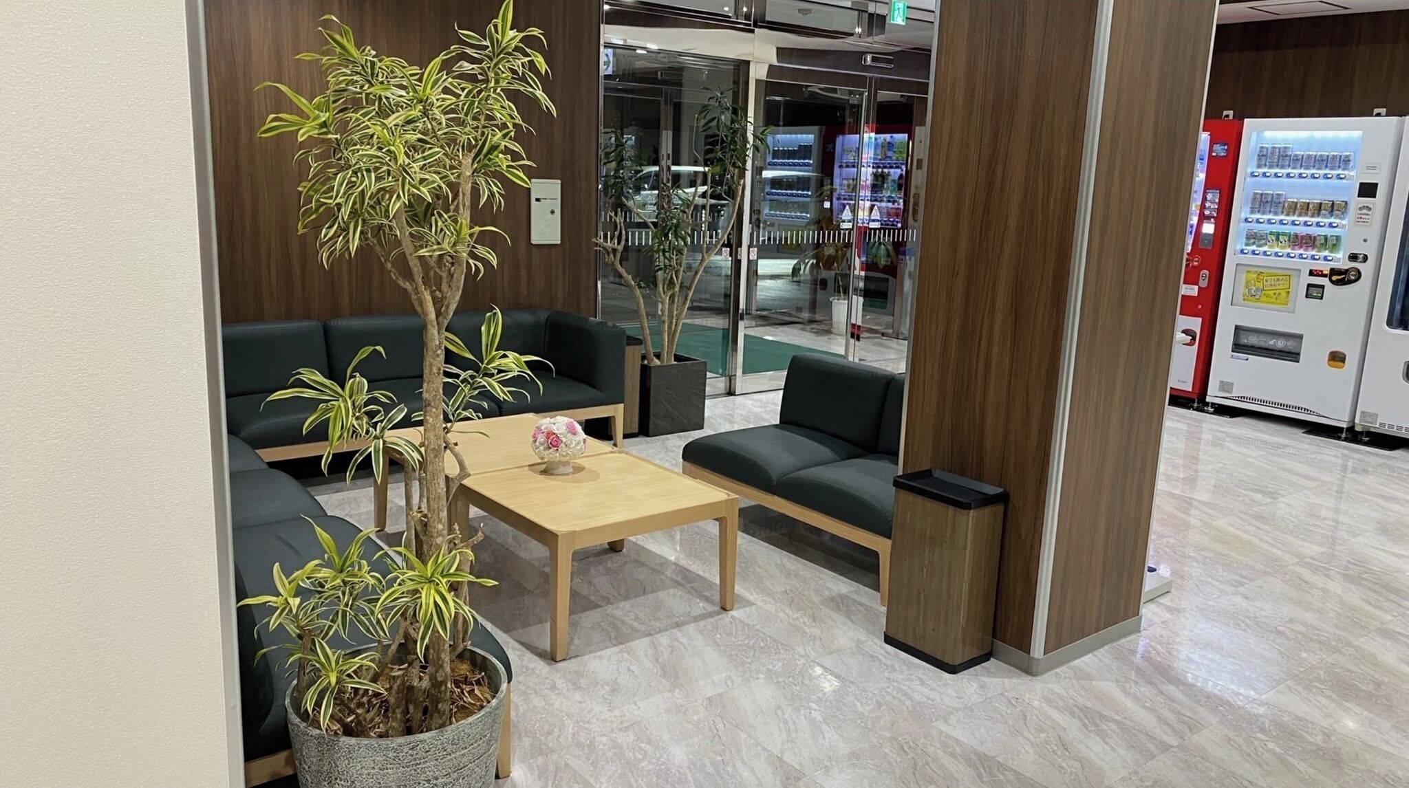 A modern lobby with dark green sofas, a wooden coffee table, and potted plants.