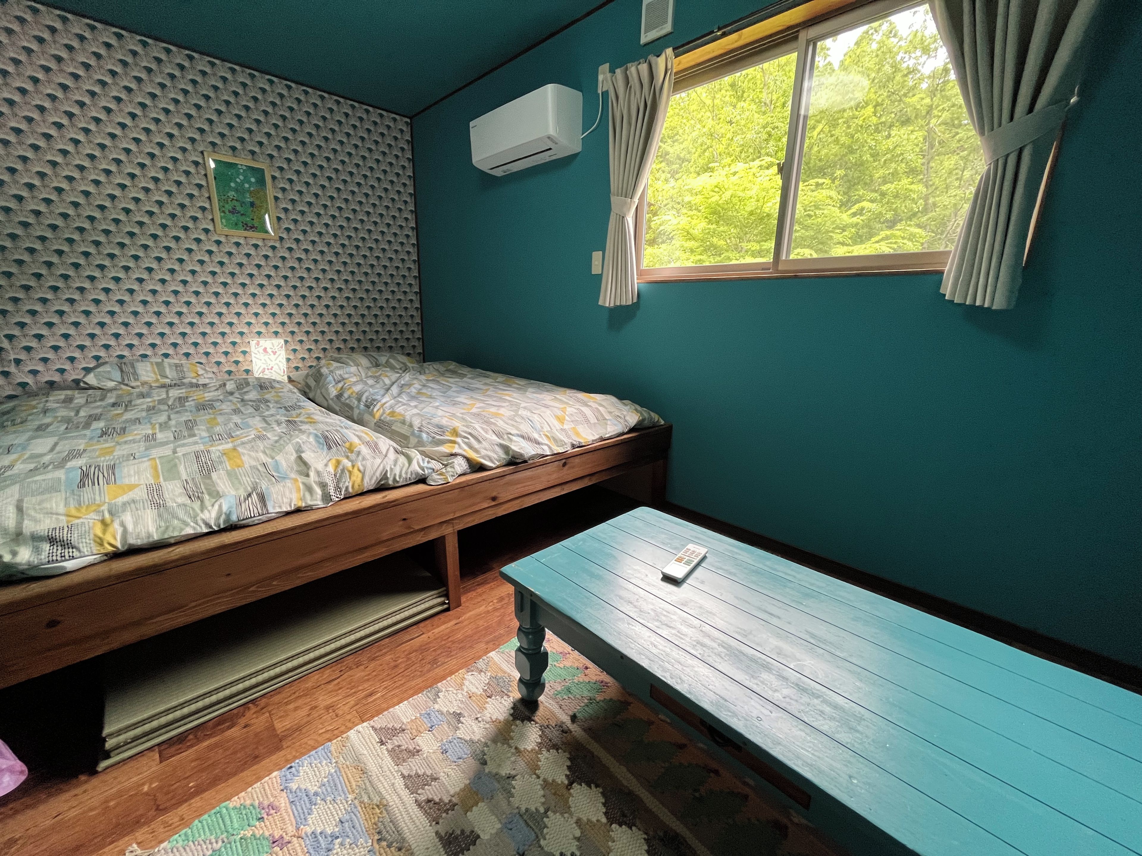A room with two beds, a window with curtains, an air conditioning unit, and a wooden bench.