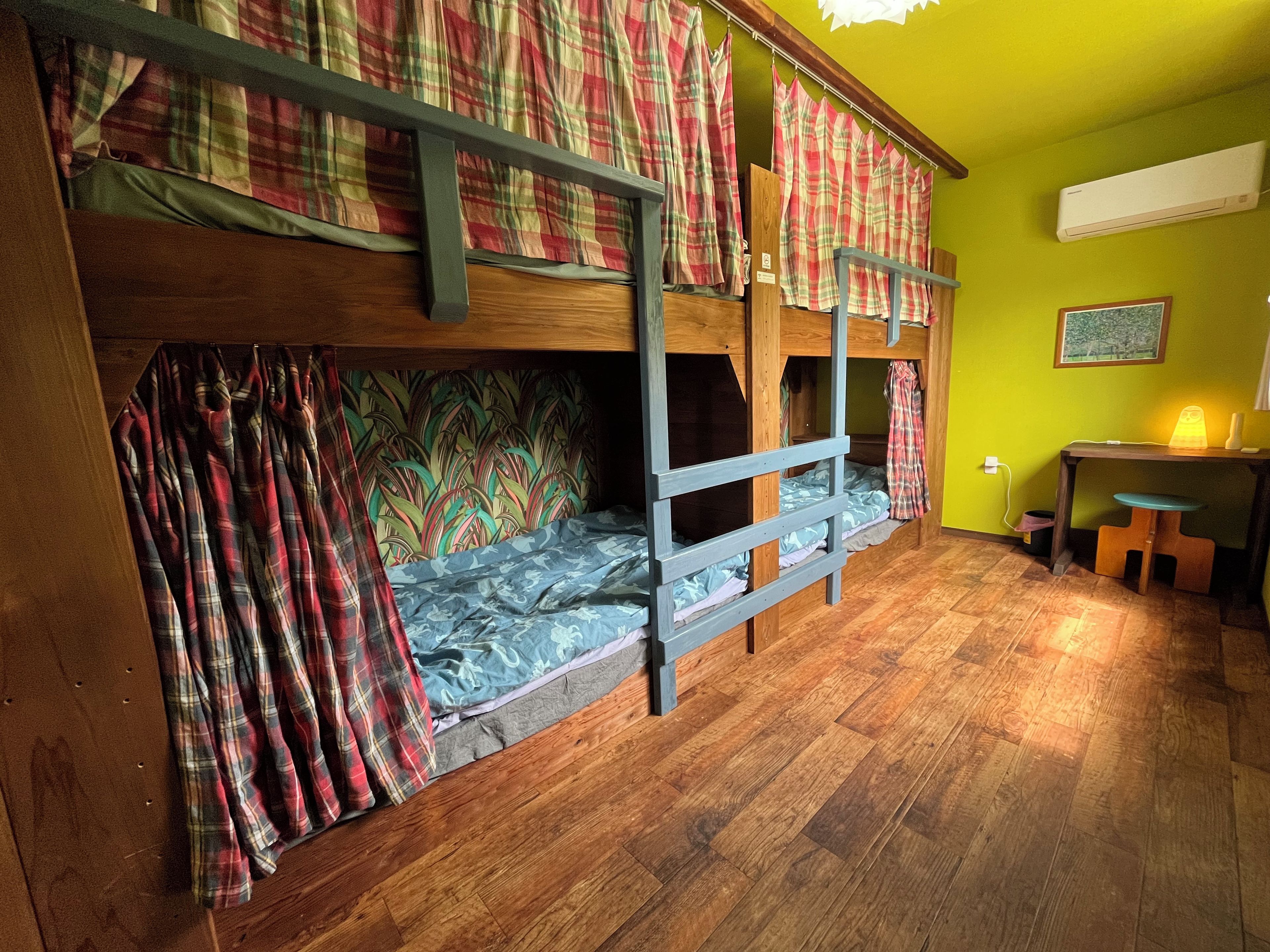 A bunk bed room with plaid curtains, a wooden floor, a desk with a chair, and an air conditioner on the wall.