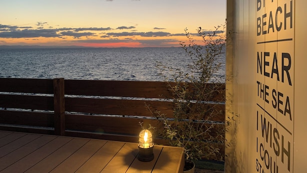 A wooden deck with a view of the sea at sunset, a hanging light fixture, and a sign reading "BY THE BEACH NEAR THE SEA I WISH TO STAY". - Isola Base Awaji (Awaji)