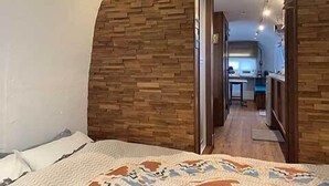 A bedroom with a bed, a wooden wall, and a hallway leading to a kitchen area. - Isola Base Awaji (Awaji)