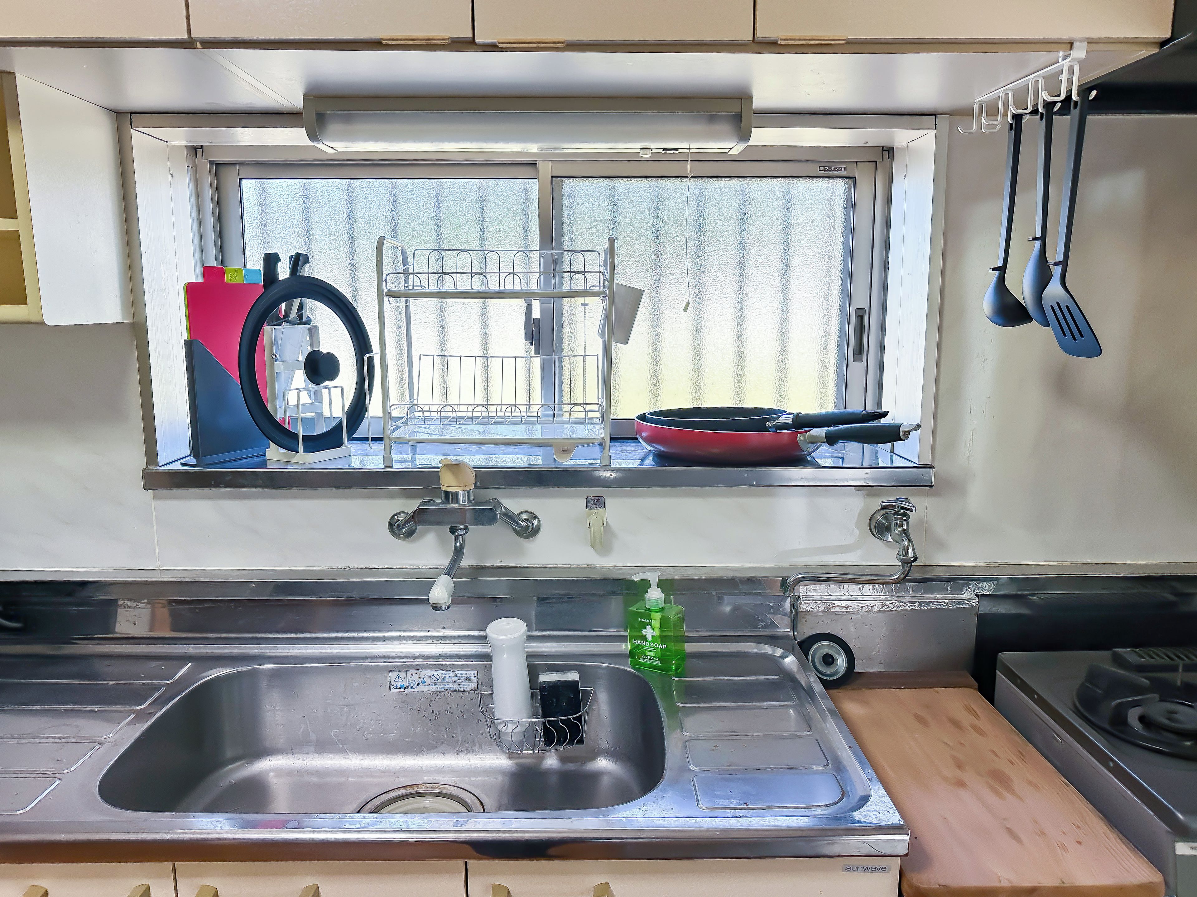 Una cocina con un lavamanos de acero inoxidable, una ventana con vistas y una sartén roja en el alféizar.