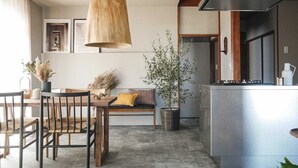 A modern kitchen with a dining area, featuring a wooden dining table, chairs, a bench, a hanging light fixture, and a potted plant. - Maigre Flat (Ito)